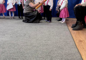 Dzieci stoją w półkolu, dziewczynki mają białe bluzki i rajstopy oraz różowe spódnice, chłopcy mają czarne spodnie i białe koszule. W tle napis pasowanie na przedszkolaka na granatowym tle. Pani dyrektor kuca przy dziecku i z pomocą dekoracyjnego ołówka dotyka ramienia dziecka