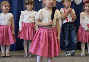 Dziewczynka stoi na środku sali z mikrofonem w ręku, ma białe rajstopy i koszulkę oraz różową spódnicę