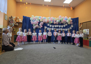 Dzieci stoją w półkolu, dziewczynki mają białe bluzki i rajstopy oraz różowe spódnice, chłopcy mają czarne spodnie i białe koszule.