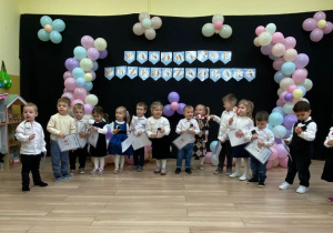 Dzieci stoją w półkolu, ubrane na galowo, w tle napis na czarnym tle, pasowanie przedszkolaka