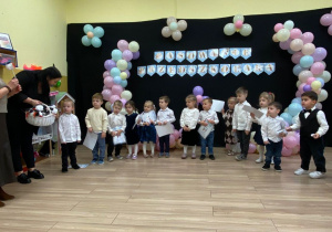 Dzieci stoją w półkolu, ubrane na galowo, w tle napis na czarnym tle, pasowanie przedszkolaka. Obok stoją dwie Panie.