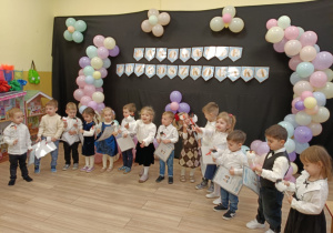 Dzieci stoją w półkolu, ubrane na galowo, w tle napis na czarnym tle, pasowanie przedszkolaka