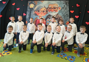 Chłopcy w białych koszulach klękają na jedno kolano przed dekoracją na dzień babci i dziadka