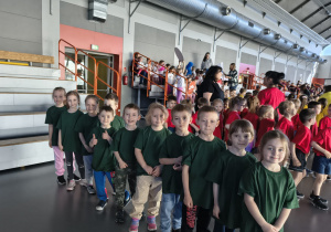 Dzieci stoją w rzędach przygotowując się do konkurencji, jedni mają zielone koszulki, inni czerwone.