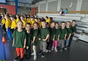 Dzieci stoją w rzędach przygotowując się do konkurencji, jedna drużyna ma zielone koszulki, druga czerwone.