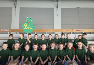 Dzieci ubrane w jednakowe, zielone koszulki siedzą na trybunach kibicując innym uczestnikom