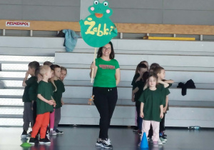 Pani trzyma proporczyk o sylwecie żabki. Jest ubrana w zieloną koszulkę i czarne spodnie.