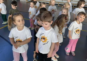 Dzieci ubrane w białe bluzki z emblematem słoneczka stoją w rozsypce