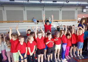 Dzieci w wraz z wychowawcą ubrani w czerwone koszulki trzymają ręce uniesione do góry.