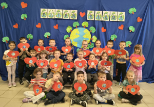 Dzieci trzymają w ręce papierowe serca i pozują do zdjęcia na tle niebieskiego materiału z symboliczną planetą ziemią oraz napisem Dzień Ziemi