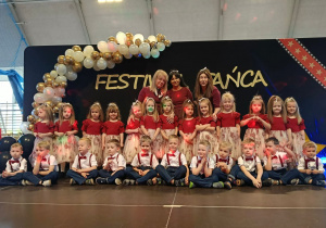 Grupa dzieci stroi w dwóch rzędach, w tle dekoracja z biało złotych balonów i napis festiwal twórczości