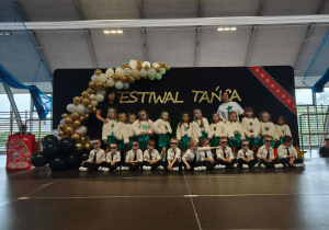 Grupa dzieci pozuje do zdjęcia, w tle dekoracja z biało złotych balonów i napis festiwal twórczości
