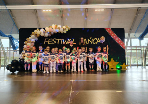Grupa dzieci pozuje do zdjęcia, w tle dekoracja z biało złotych balonów i napis festiwal twórczości