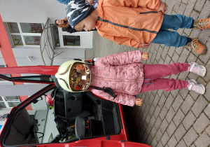 Dziewczynka w różowej kurtce zakłada kask hełm strażacki