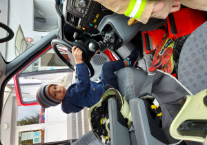 chłopiec w niebieskiej kurtce siedzi za kierownicą wozu strażackiego