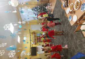 Dzieci ubrane w czerwone stroje z czapkami Mikołaja na głowie stoją w kole a po środku siedzi na krześle Mikołaj i trzyma worek pełen prezentów