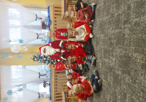 Dzieci siedzą w rozsypce pod choinką a po środku siedzi Mikołaj