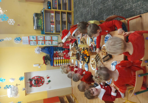 Dzieci ubrane w stroje w kolorze czerwonym siedzą przy stole, na którym rozłożone są słodycze i owoce