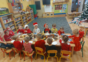 Dzieci ubrane w stroje w kolorze czerwonym siedzą przy stole, na którym rozłożone są słodycze i owoce