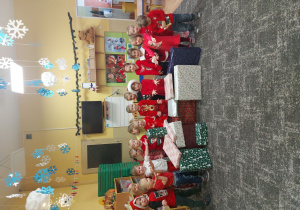 Dzieci siedzą na środku dywanu pokazując na duże prezenty jakie dostały. Prezenty owinięte są w świąteczny kolorowy papier