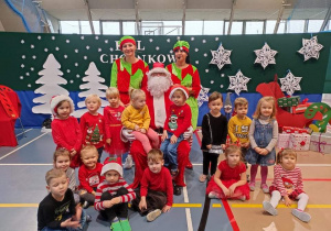 Grupa małych dzieci, ubranych w kolorowe, świąteczne stroje siedzi z Mikołajem i dwoma elfami