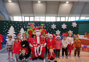 Grupa małych dzieci, ubranych w kolorowe, świąteczne stroje siedzi z Mikołajem i dwoma elfami