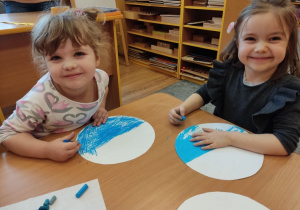 Dwie uśmiechnięte dziewczynki siedzą przy stoliku i malują pastelami.