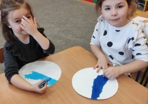 Dwie dziewczynki siedzą przy stoliku i malują pastelami.