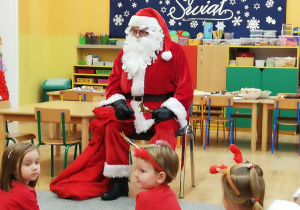 Św. Mikołaj siedzi na krześle i rozdaje prezenty dzieciom.