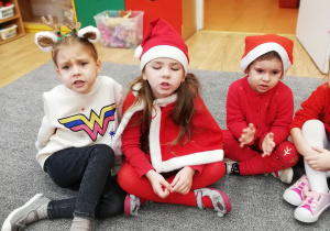 Trzy dziewczynki ubrane na czerwono siedzą na dywanie i śpiewają piosenkę.