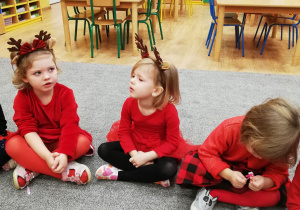Trzy dziewczynki ubrane na czerwono siedzą na dywanie i śpiewają piosenkę.