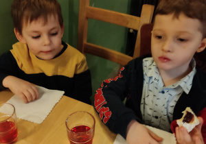 Dwóch chłopców siedzi przy stolikach i jedzą ciasteczko.