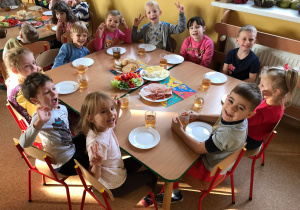 Grupa dzieci uśmiecha się do zdjęcia, na stole przygotowane są półmiski ze zdrowym śniadankiem.
