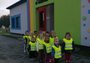 Dzieci w kamizelkach odblaskowych stoją w parach przed przedszkolem.