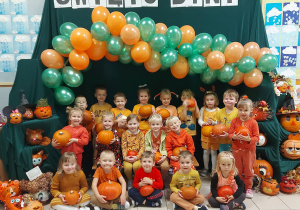 Dzieci stoją pod girlandą z pomarańczowo, zielonych balonów, trzymają w ręce duże pomarańczowe dynie. Nad nimi napis Święto Dyni