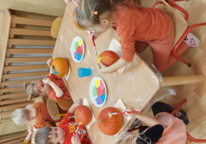Dzieci w kilkuosobowych grupach przy stolikach dekorują swoje dynie kolorowymi farbami