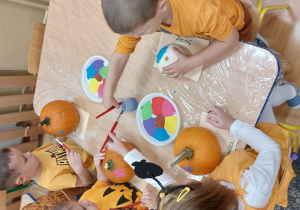 Dzieci w kilkuosobowych grupach przy stolikach dekorują swoje dynie kolorowymi farbami