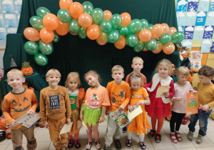 Dzieci ubrane na pomarańczowo stoją pod girlandą z pomarańczowo zielonych balonów i pozują do zdjęcia