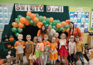 Dzieci ubrane na pomarańczowo stoją pod girlandą z pomarańczowo zielonych balonów i pozują do zdjęcia
