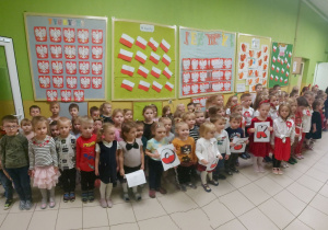 Dzieci stoją w dużej grupie, trzymają w ręce napis POLSKA, małe papierowe flagi naszego kraju oraz śpiewają hymn Polski