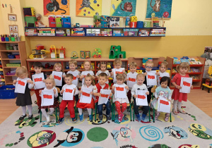 Dzieci stoją w dużej grupie, trzymają w ręce napis POLSKA, małe papierowe flagi naszego kraju oraz śpiewają hymn Polski