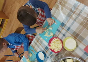 Dzieci siedzą przy stoliku i kroją jabłka na szarlotkę