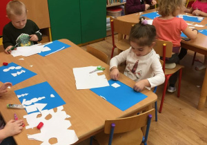 Dziewczynka i chłopiec siedzi przy stolikach i przykleja elementy z białych kartek.
